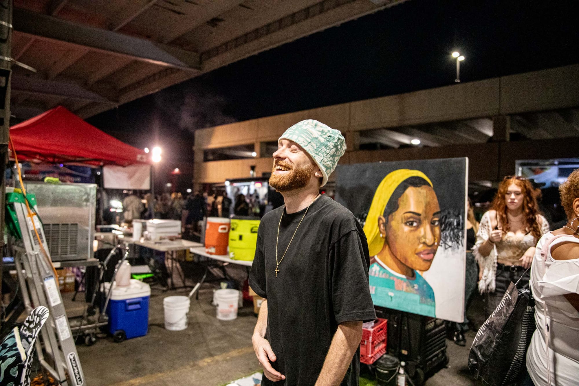 A man in a black shirt and a green and white beanie stands smiling in front of a painting of a woman in a yellow beanie. 