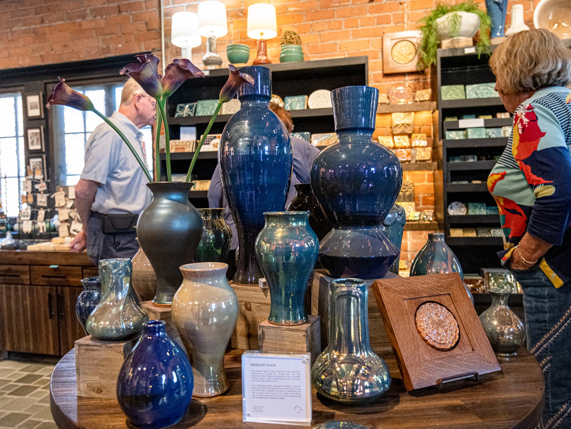 A blue coated iridescent vase stands on a table in the center of a room. Shelves of ceramics are in the background.