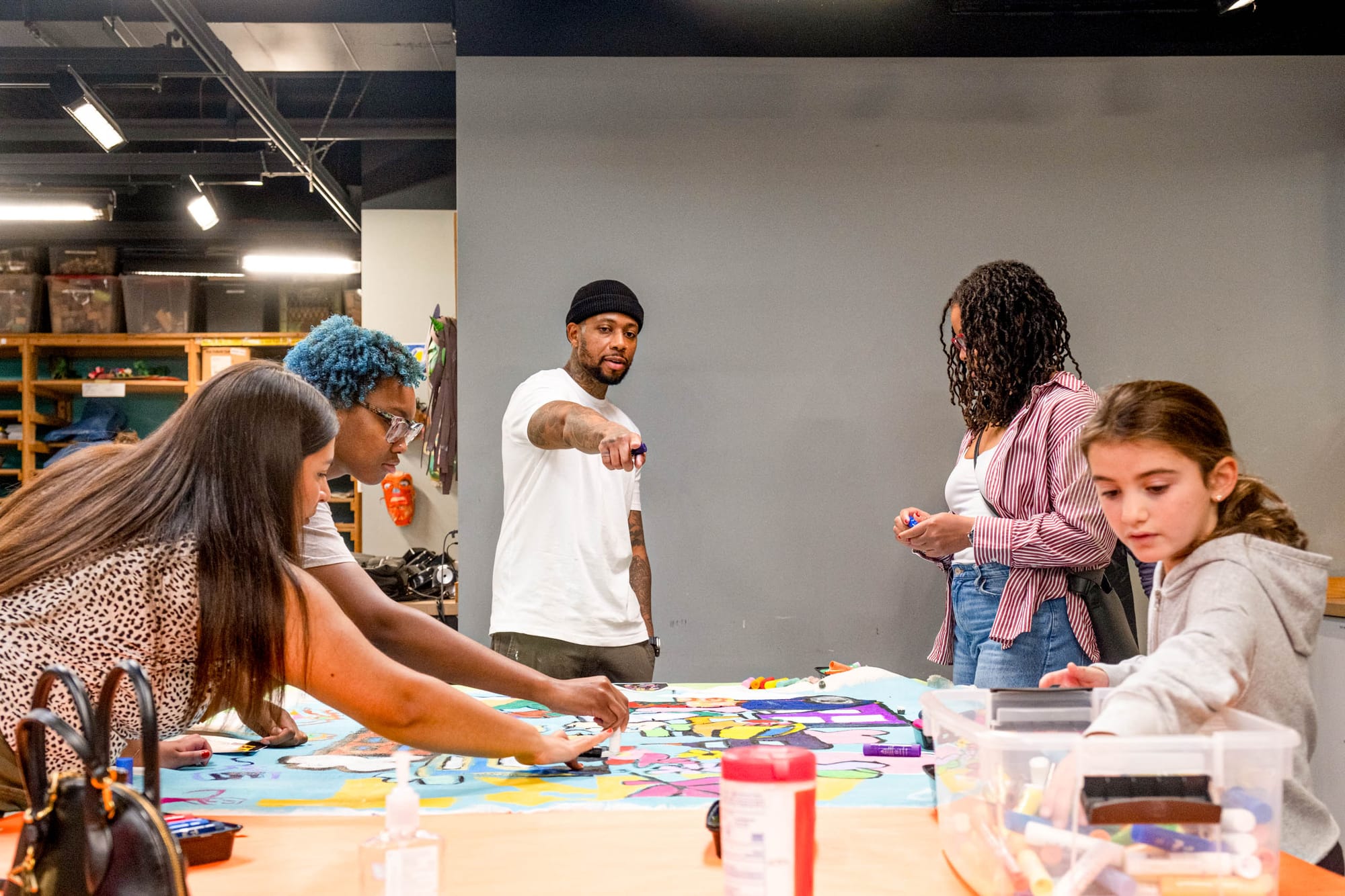 A man wearing a white shirt stands in the center frame pointing at the table. A group of four people listen and draw beside him. 