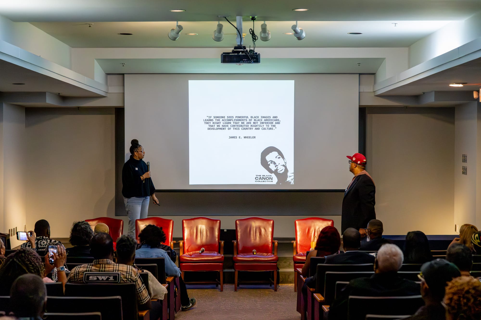 Two people stand on opposite sides of a projector screen with microphones, as the audience watches and listens in the auditorium.
