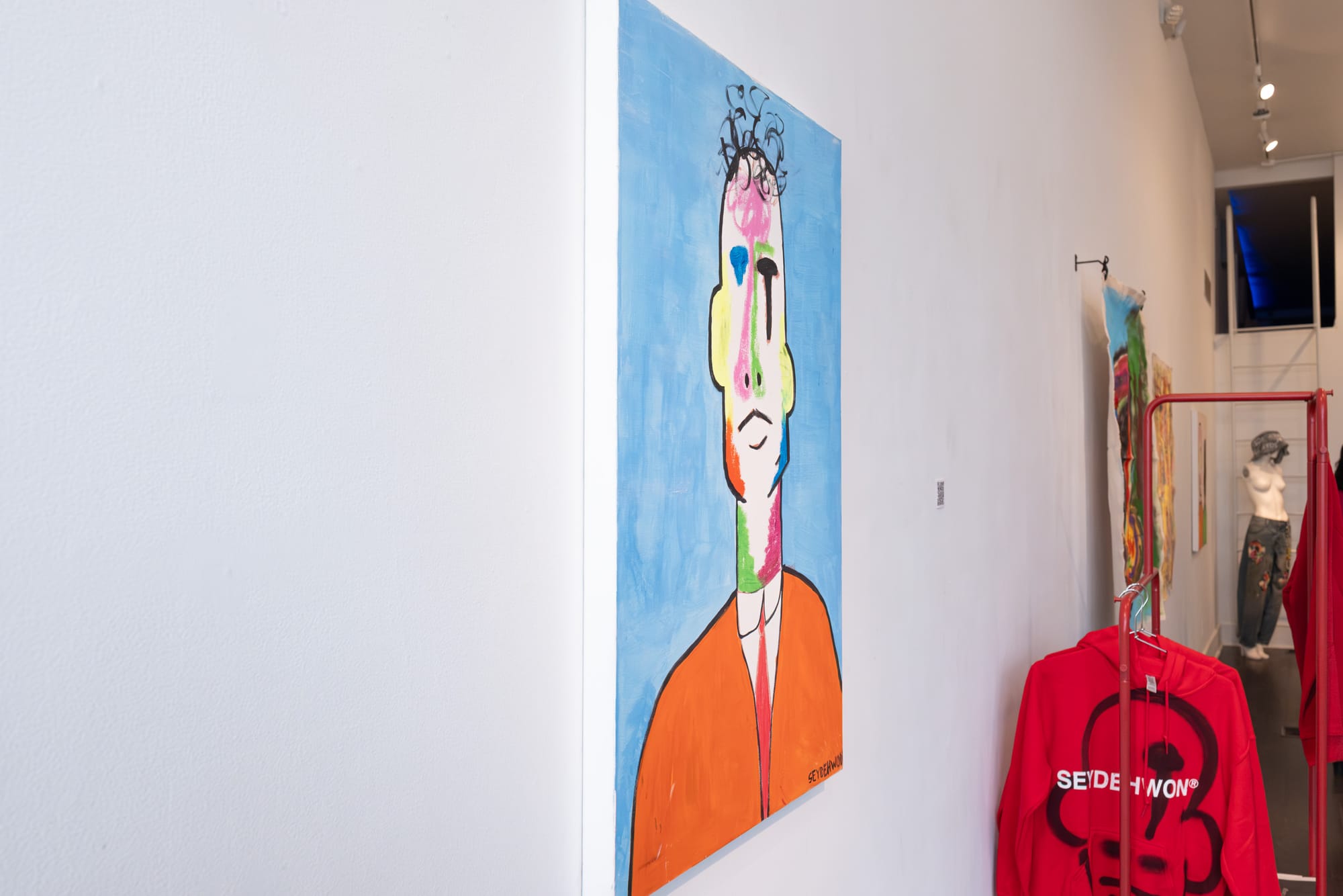 Two people stand before vibrant abstract paintings, observing a mannequin dressed in a jean jacket.