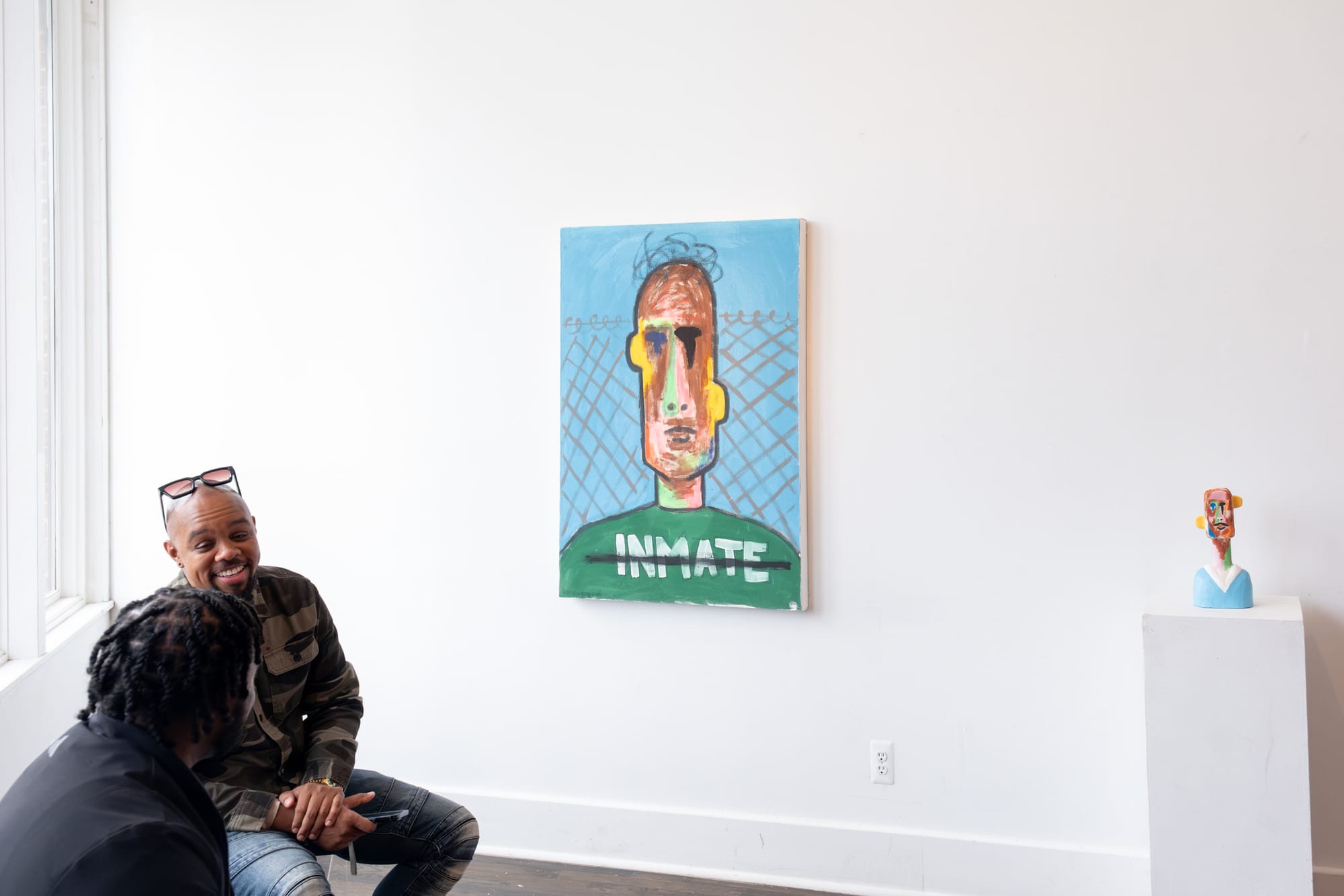 Two people sit down talking — one has glasses on his head. In the background, there is a vibrant abstract painting and sculpture against a white wall.