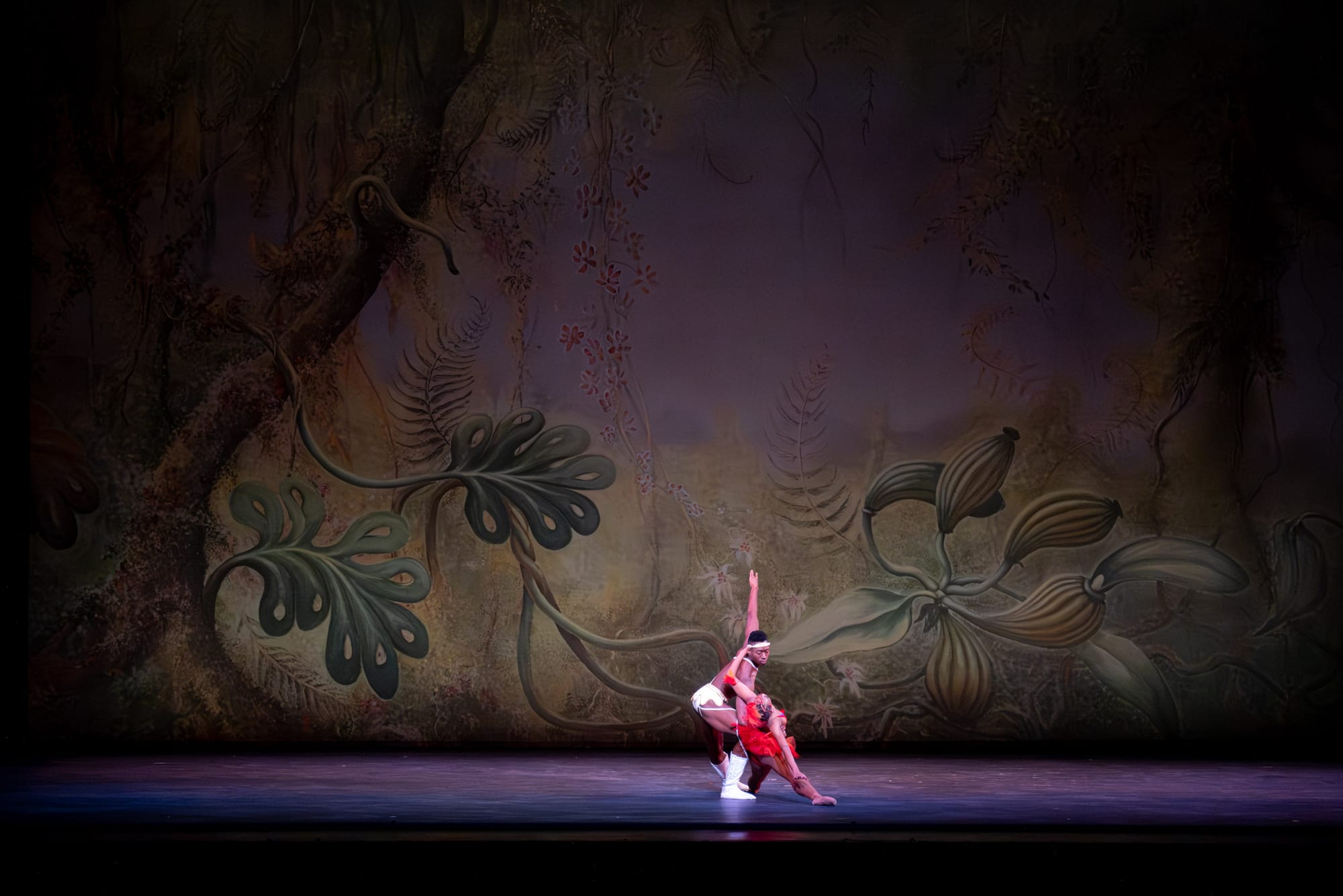 Two dancers stand on stage, one dressed in all white and the other in red. They hold each other as the dancer in red drops into a split.