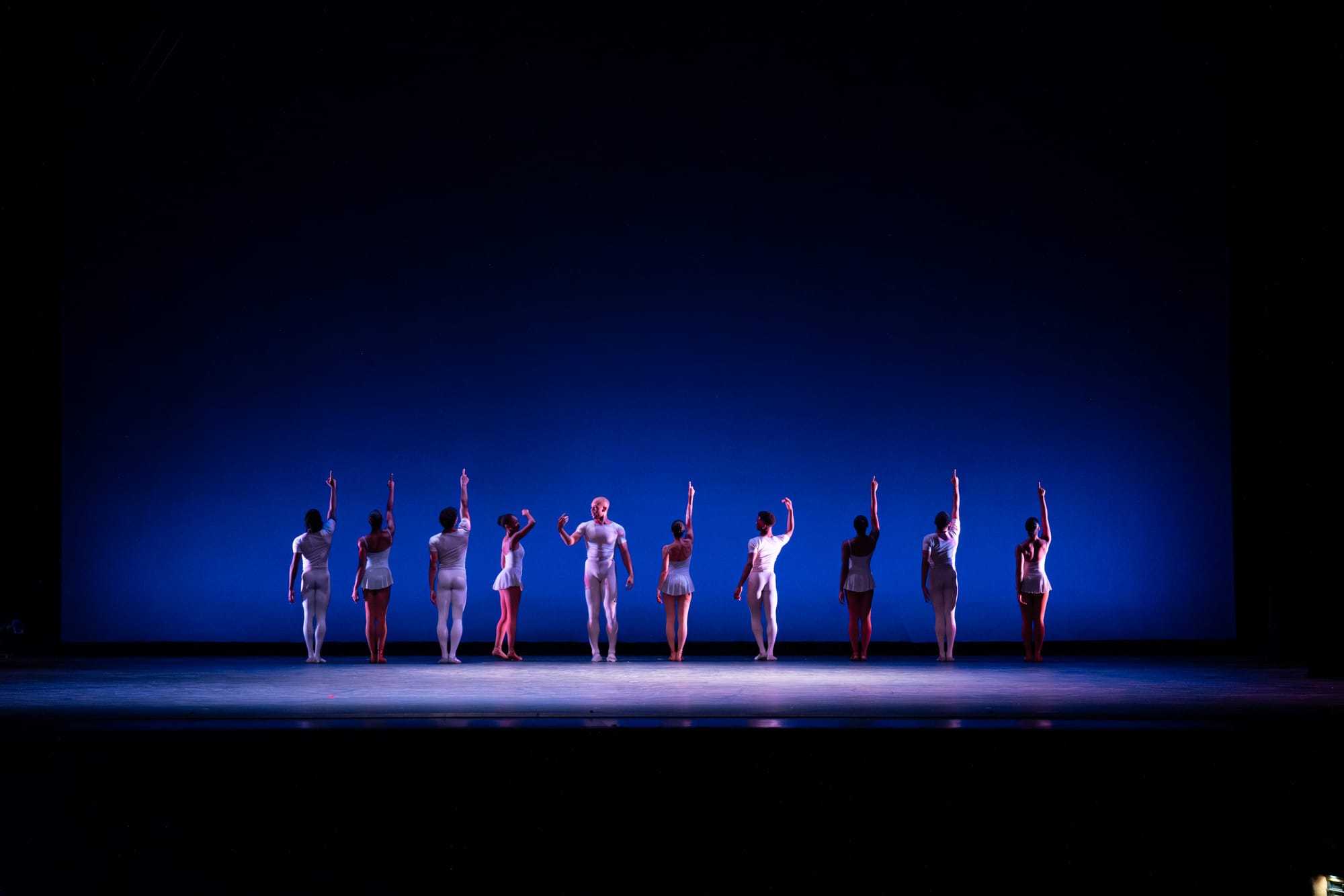 A group of people stand on a stage in the background. There is a blue background some members point at. 