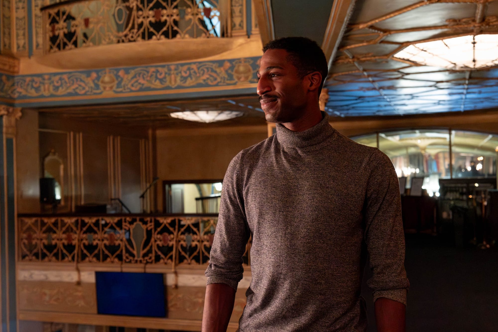 A man stands smiling, holding onto the staircase railing in a golden foyer.