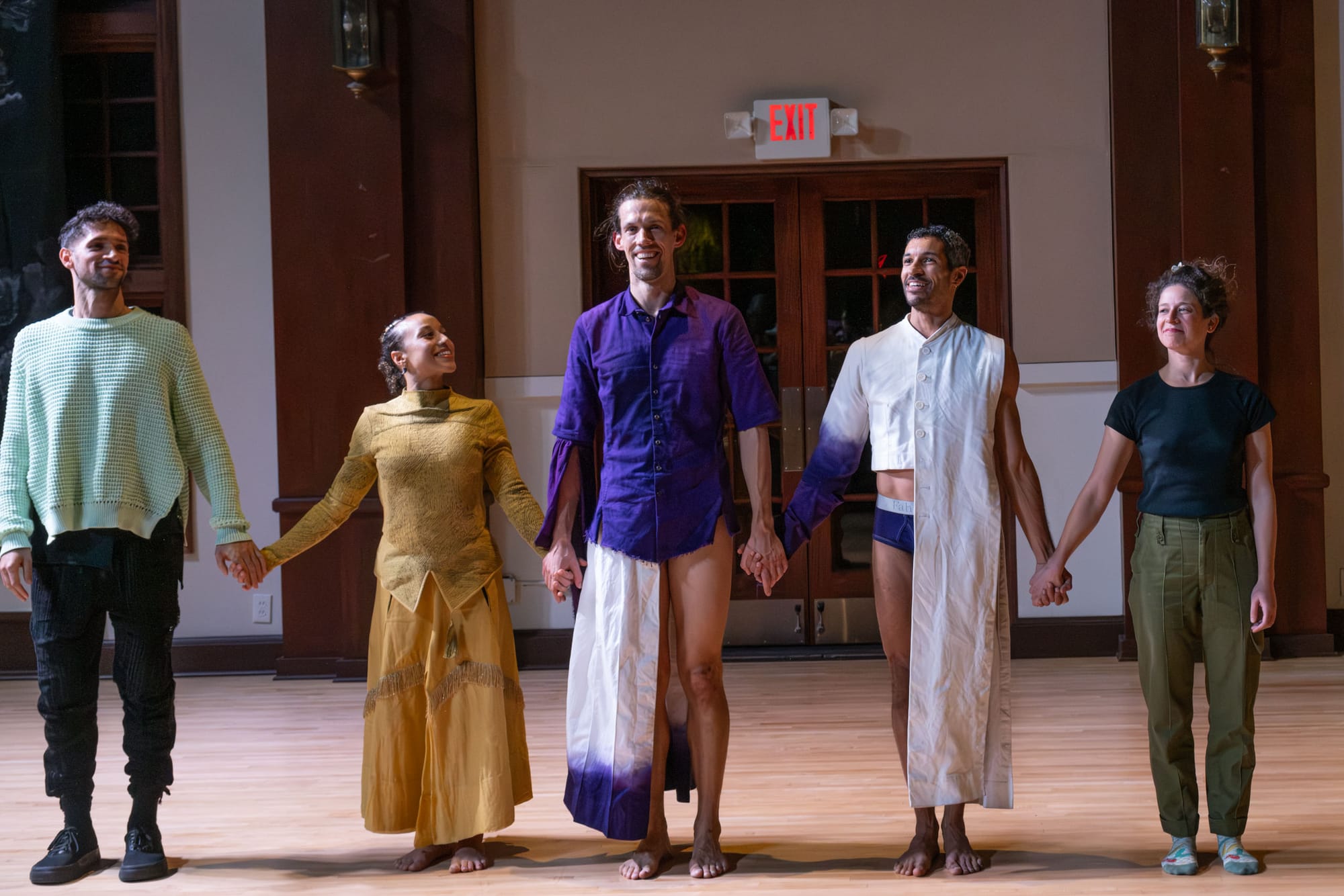 A group of people stand in the center of a room holding hands in front of an audience. 