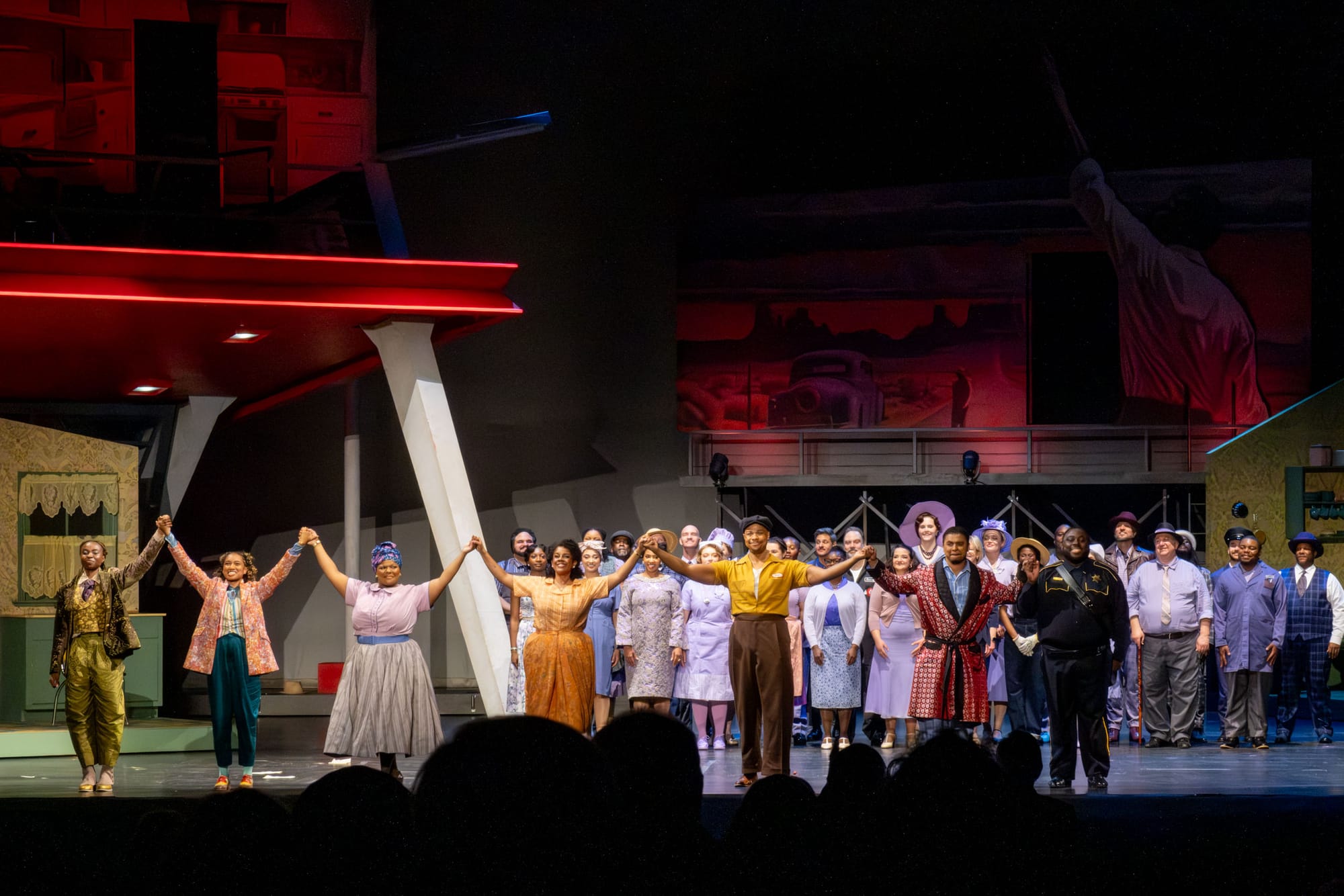 A group of performers stand on a stage with arms raised, bowing to an audience.