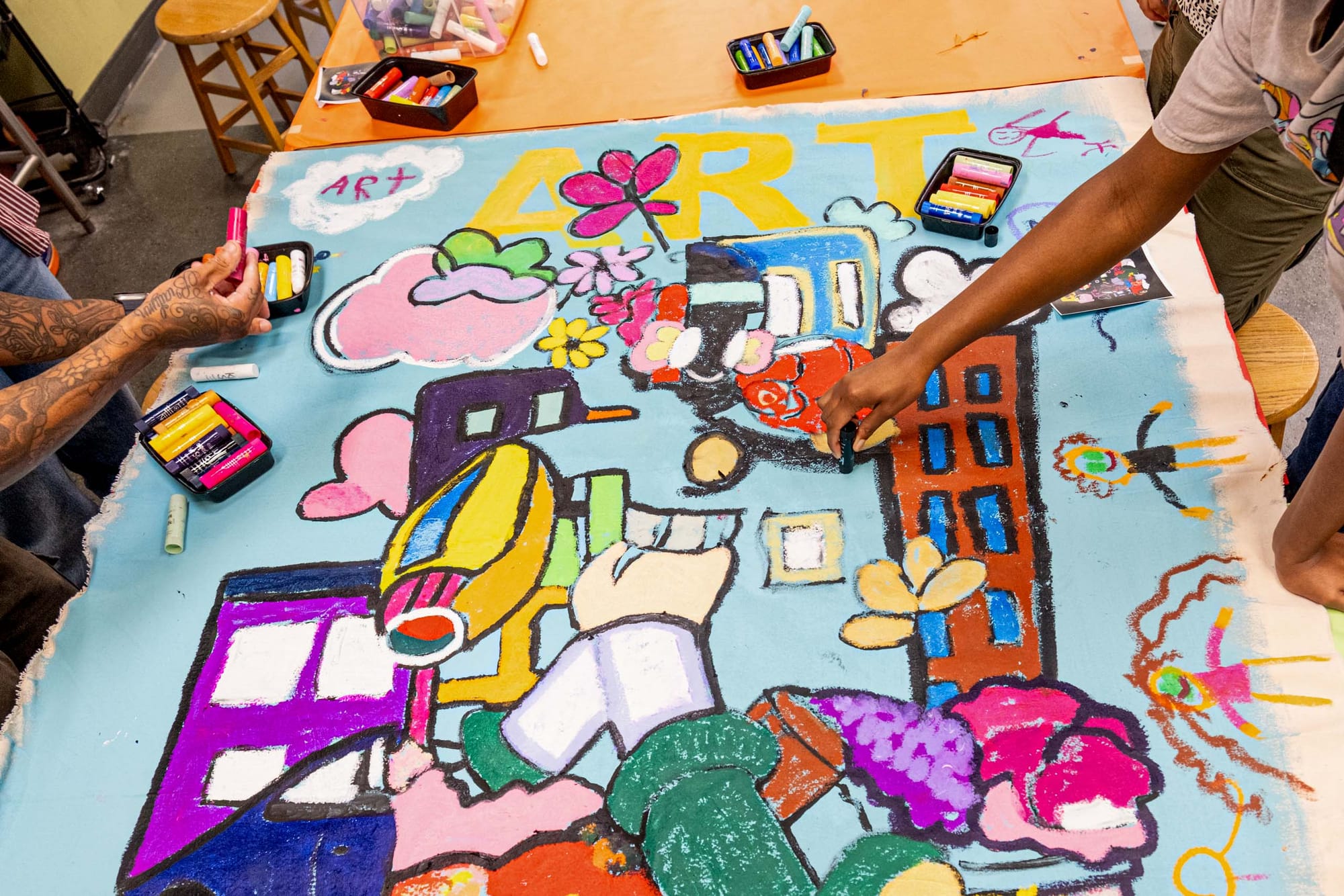 A piece of artwork lies on the table, arms reach out and contribute to the painting using pieces of charcoal.