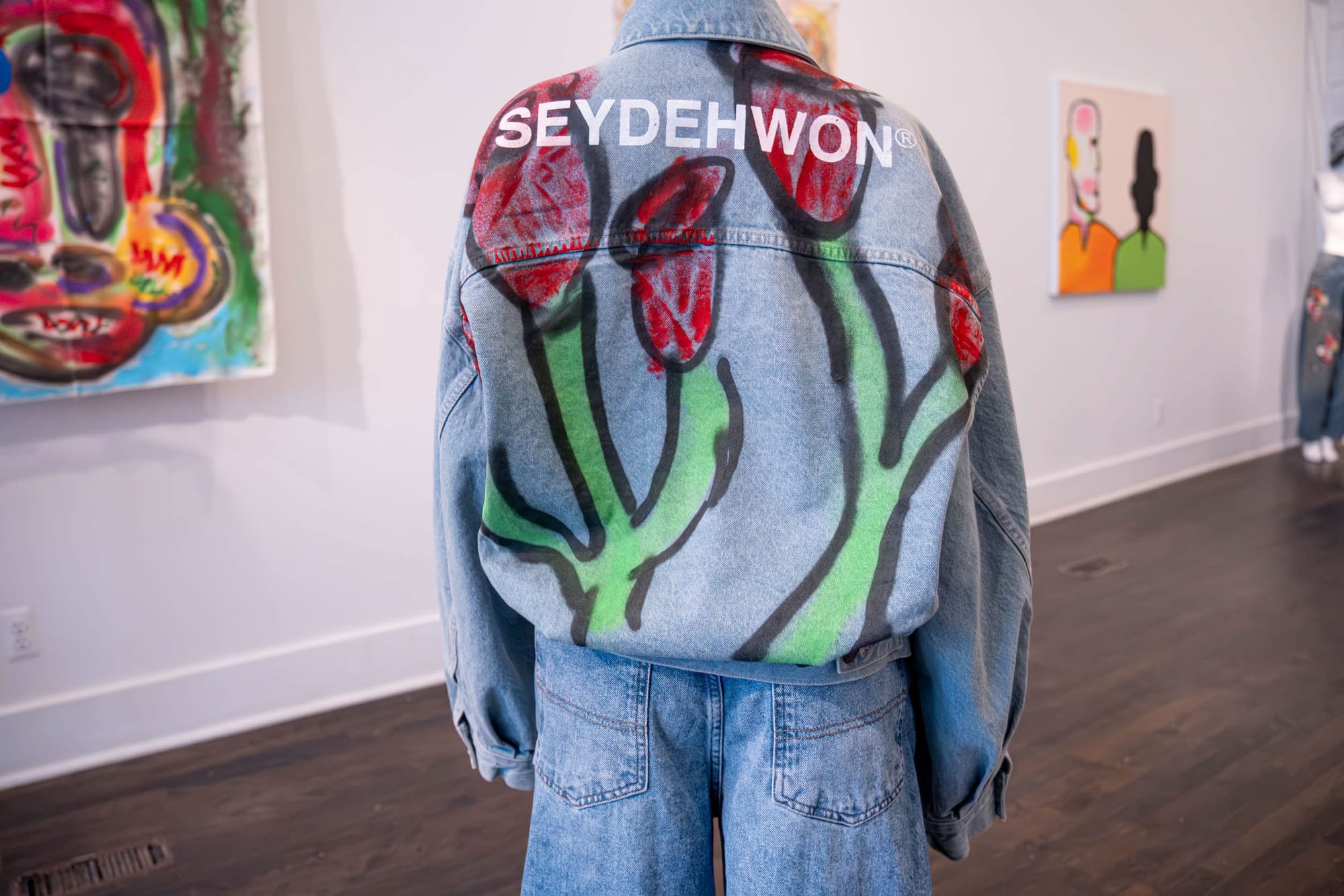 A mannequin wearing a denim set embroidered with roses and “Seydehwon” across the back stands in the middle of the gallery, framed by paintings in the background.