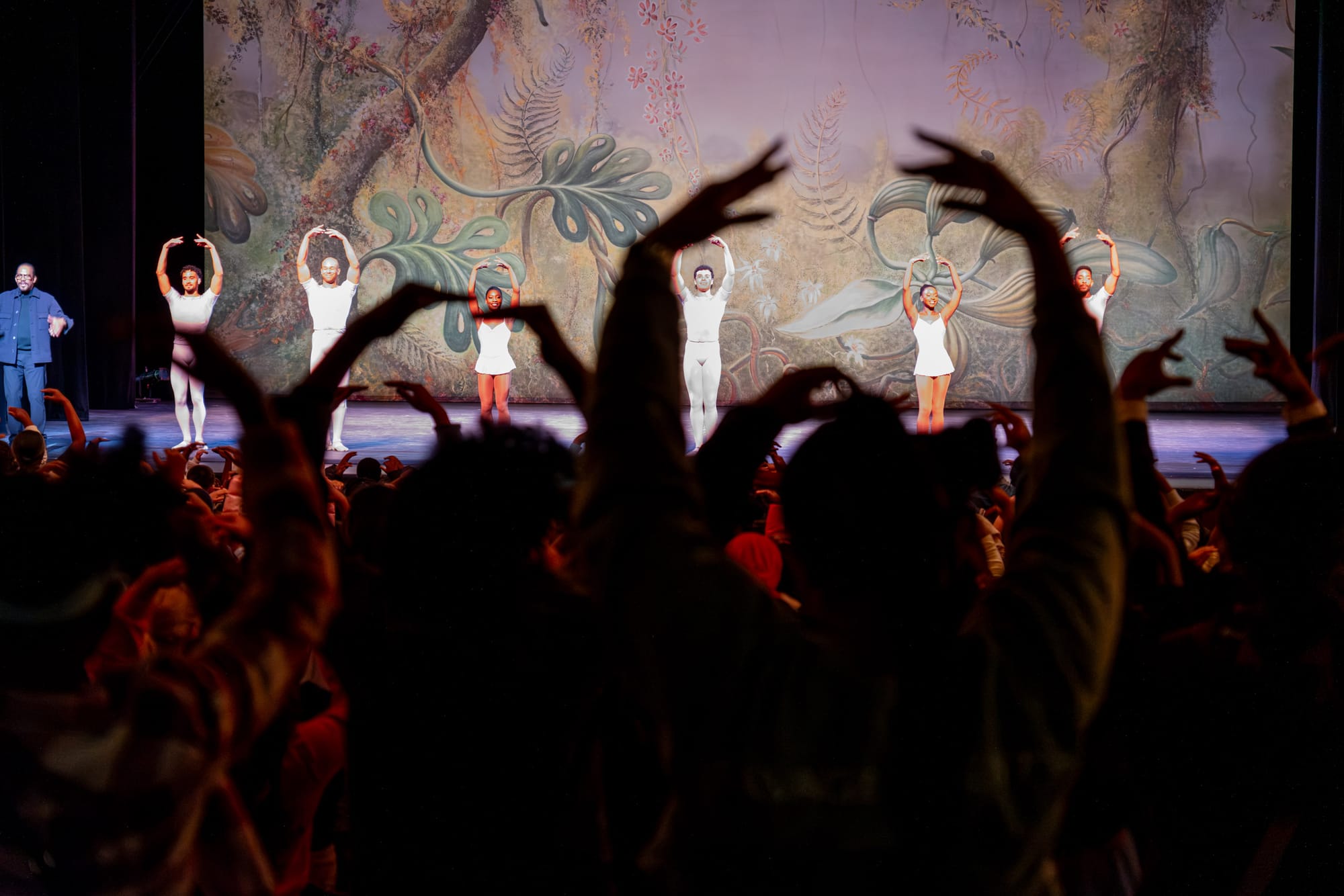 An audience of people hold their hands up while people on stage in front do the same. There is a nature background displayed on a screen behind them.  