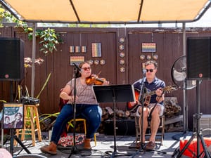 Two musicians sit on a stage under a tent, one playing the violin and the other playing the guitar.