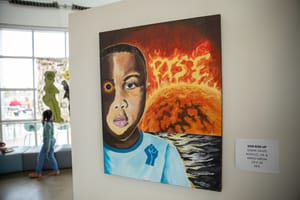A painting of a young Black boy with a raised fist on his shirt, one eye glowing orange, stands before a fiery sunrise with the word "RISE" in the sky, displayed in a gallery.