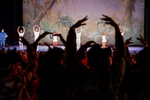 An audience of people hold their hands up while people on stage in front do the same. There is a nature background displayed on a screen behind them.  